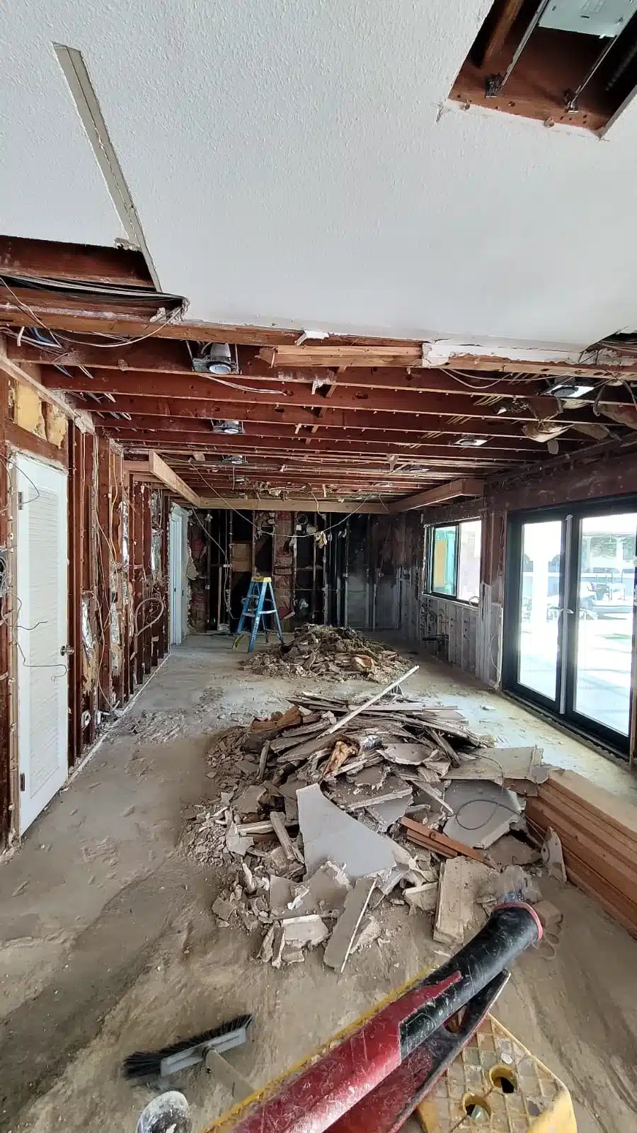 Stripped interior with exposed wall studs, ceiling joists, wiring, a blue ladder and piles of demolition debris.