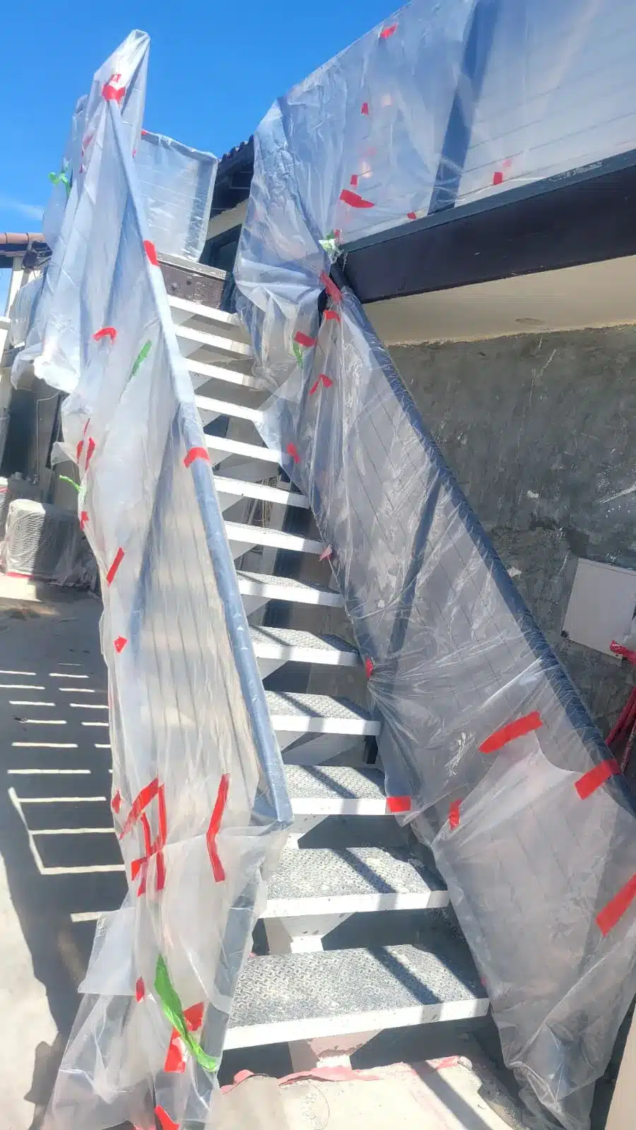 Exterior metal staircase with railings covered in clear plastic sheeting secured with red and green tape.