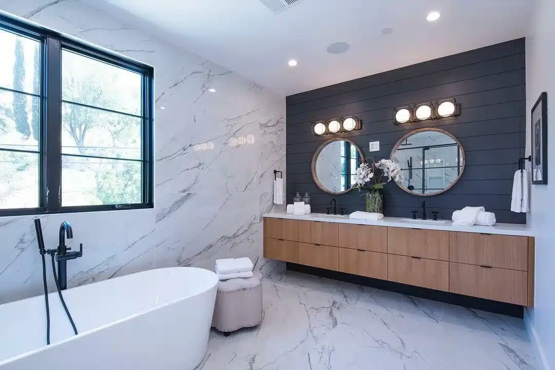 Finished bathroom with marble-look surfaces, a white freestanding tub, wood vanity, round mirrors, and a dark