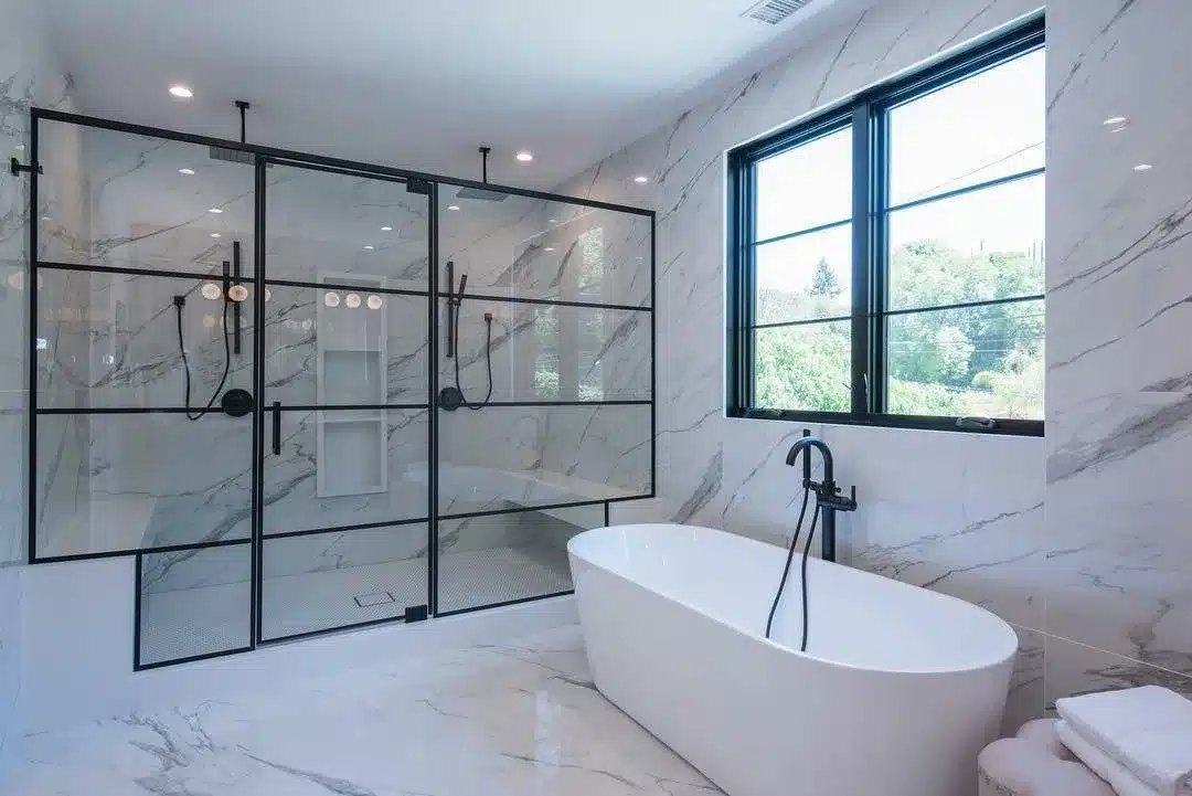 Finished bathroom with a freestanding white tub beside a black-framed glass shower, marble-look walls, and a large window.