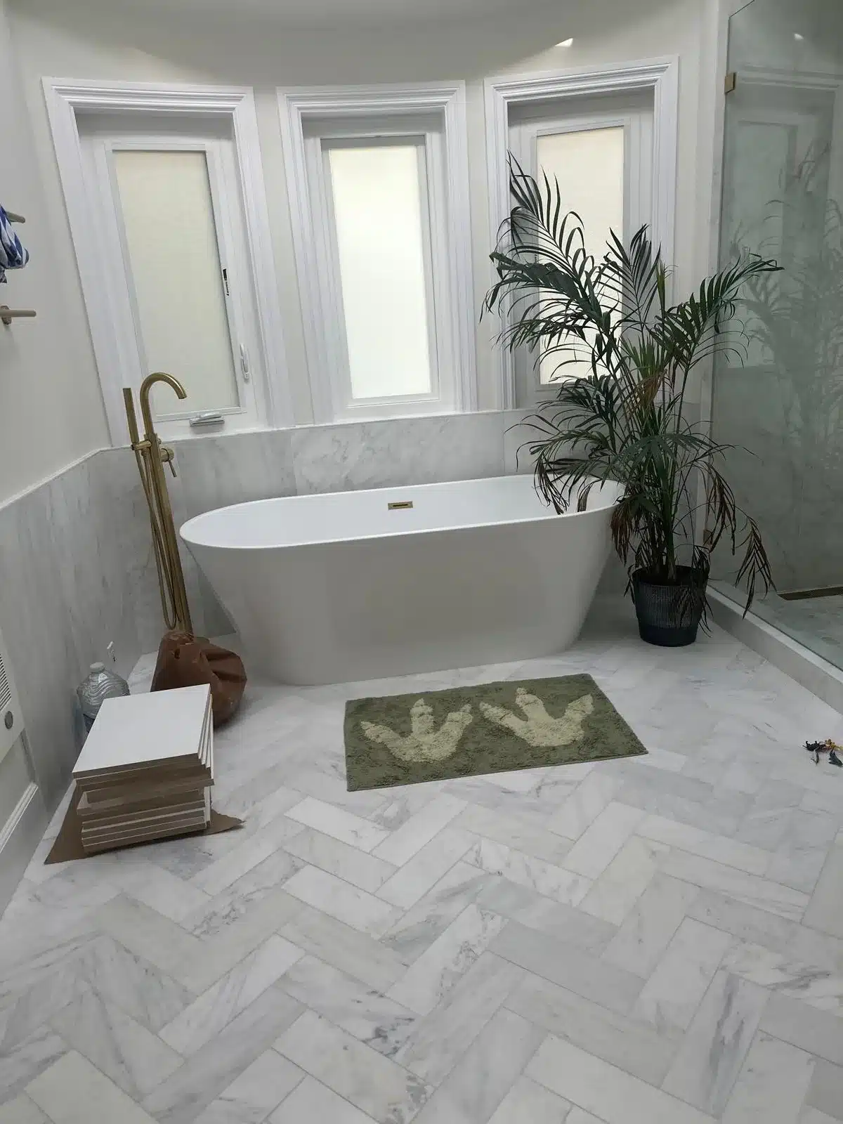 Finished tub area with white freestanding tub, brass-colored floor-mounted filler, shaded windows, and herringbone marble