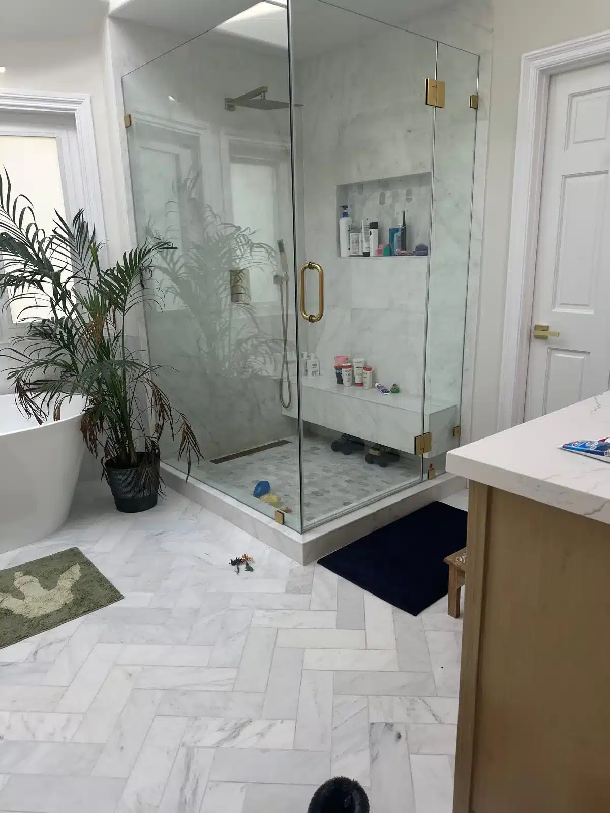 Finished glass-enclosed marble shower with recessed niche, bench, brass-colored hardware, and herringbone flooring outside.