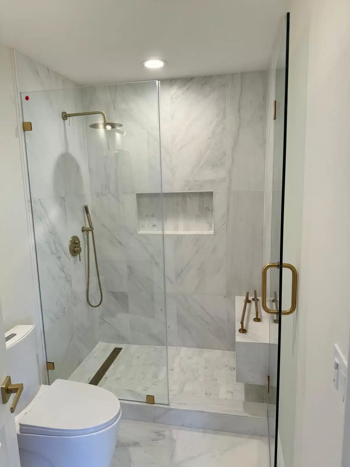 Finished marble-tiled shower with rainfall showerhead, handheld sprayer, recessed niche, linear drain, and frameless glass.