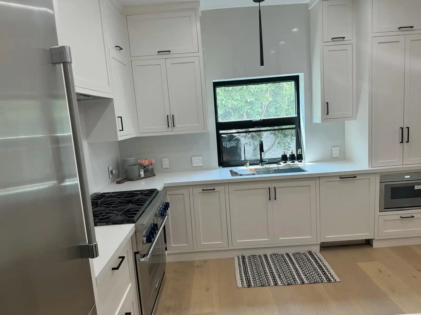 Finished kitchen with white cabinetry, black-framed sink window, pale counters, and stainless steel appliances.