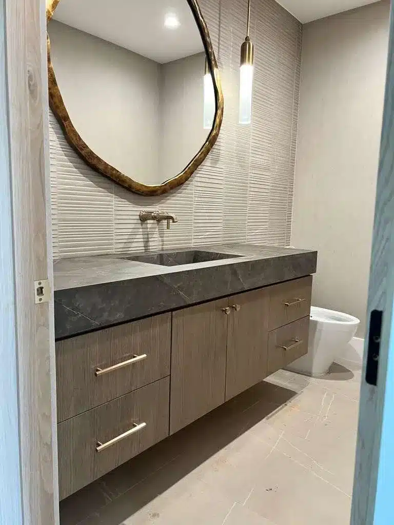 Finished bathroom vanity area with wood-grain cabinets, a dark veined countertop, rounded mirror, and pendant lighting.