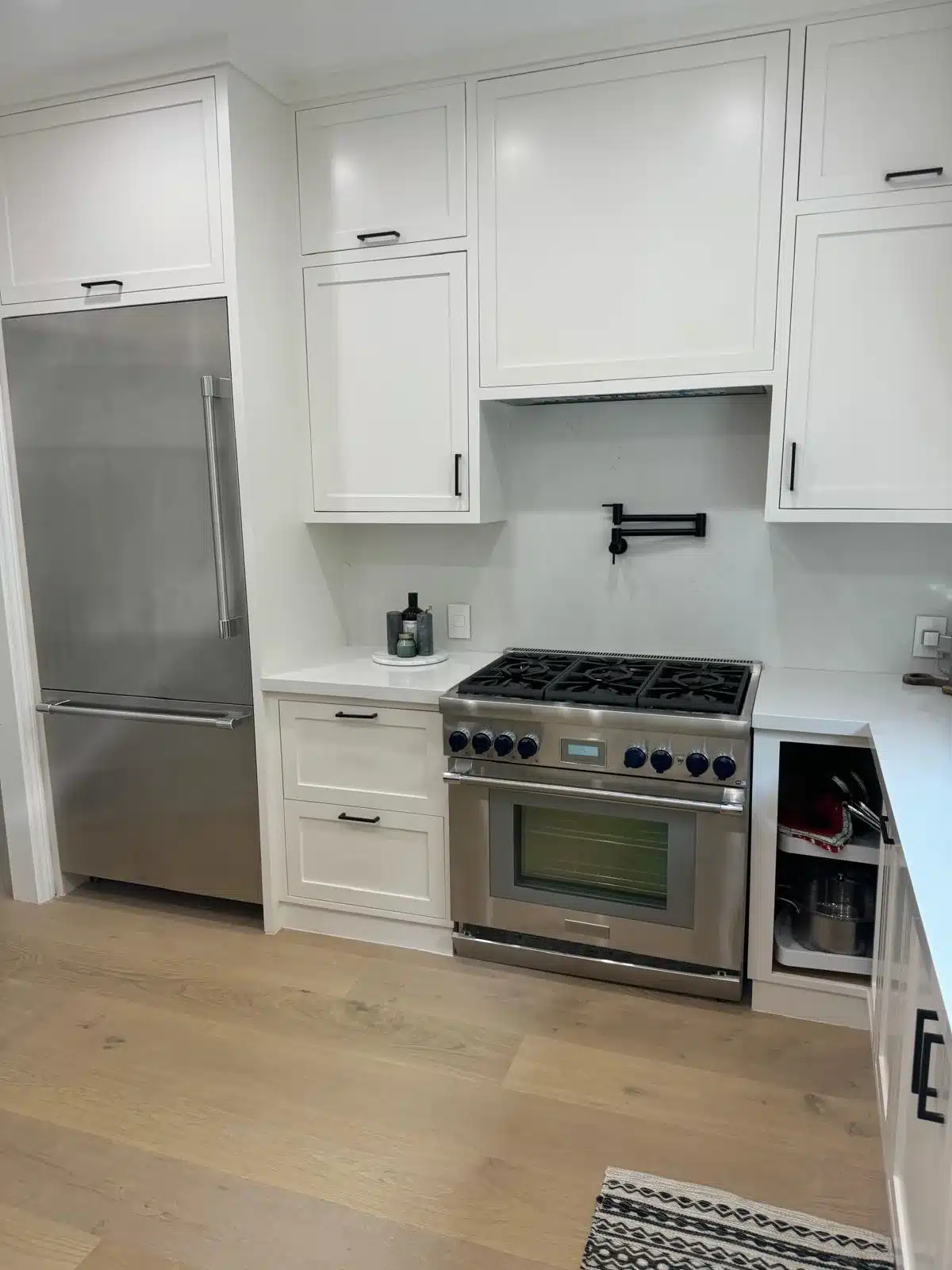 Finished kitchen appliance wall with white paneled hood enclosure, black pot filler, and cookware in an open corner cabinet.