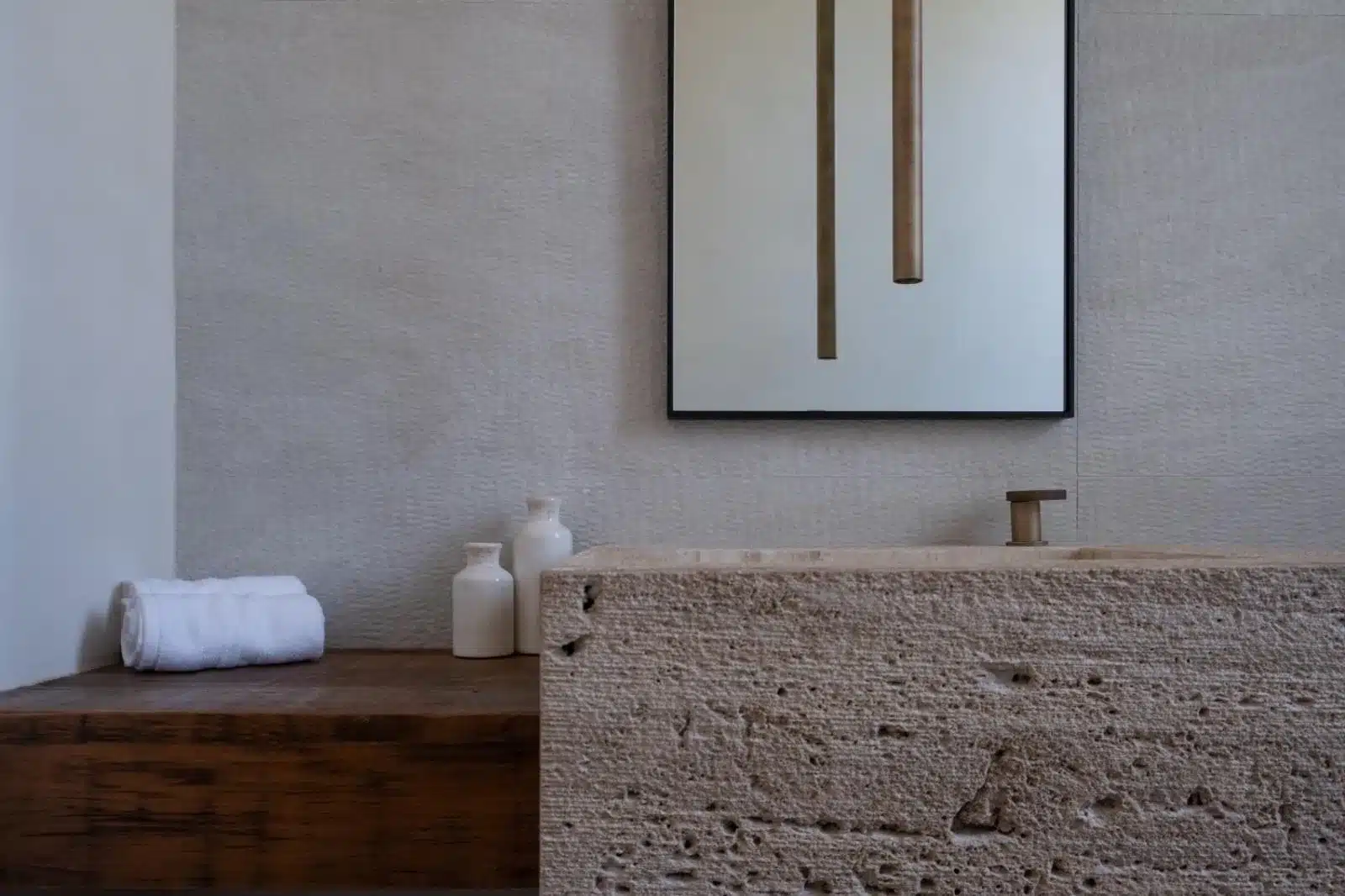 Finished bathroom detail with a rough-textured stone sink, warm metallic faucet, wood shelf, and black-framed mirror