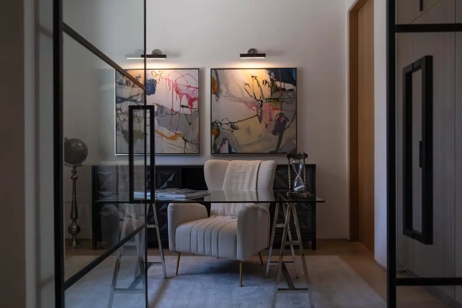 Finished office behind black-framed glass doors with a glass-top desk, cream chair, abstract artwork, and picture lights