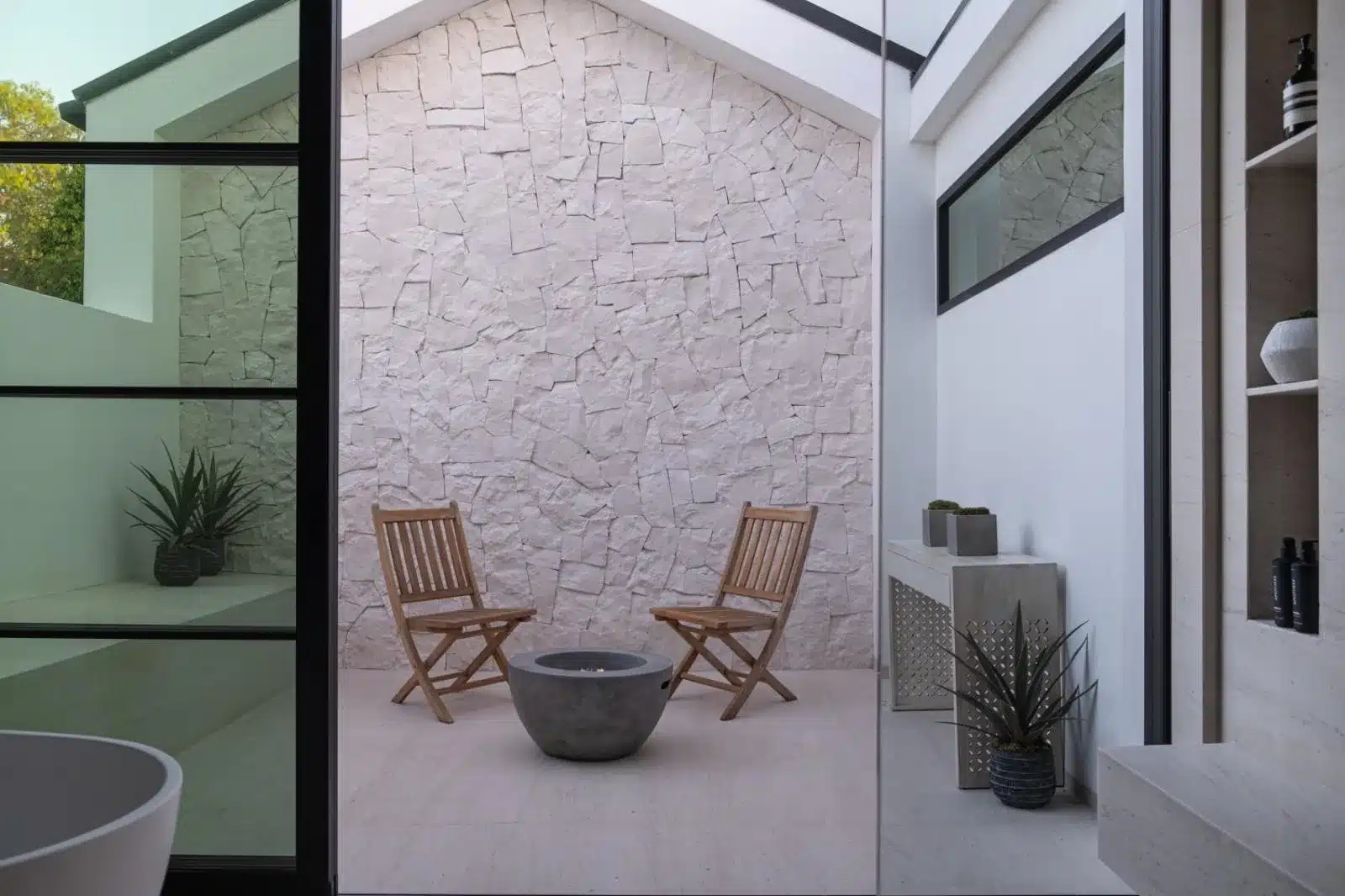 Finished courtyard with textured pale cladding, wooden folding chairs, a round fire bowl, and black-framed glazing