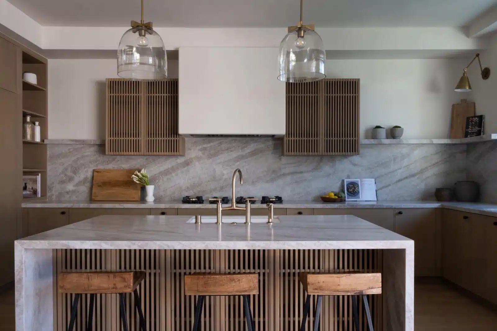 Finished kitchen with a veined waterfall island, slatted wood cabinetry, stools, and clear glass pendant lights