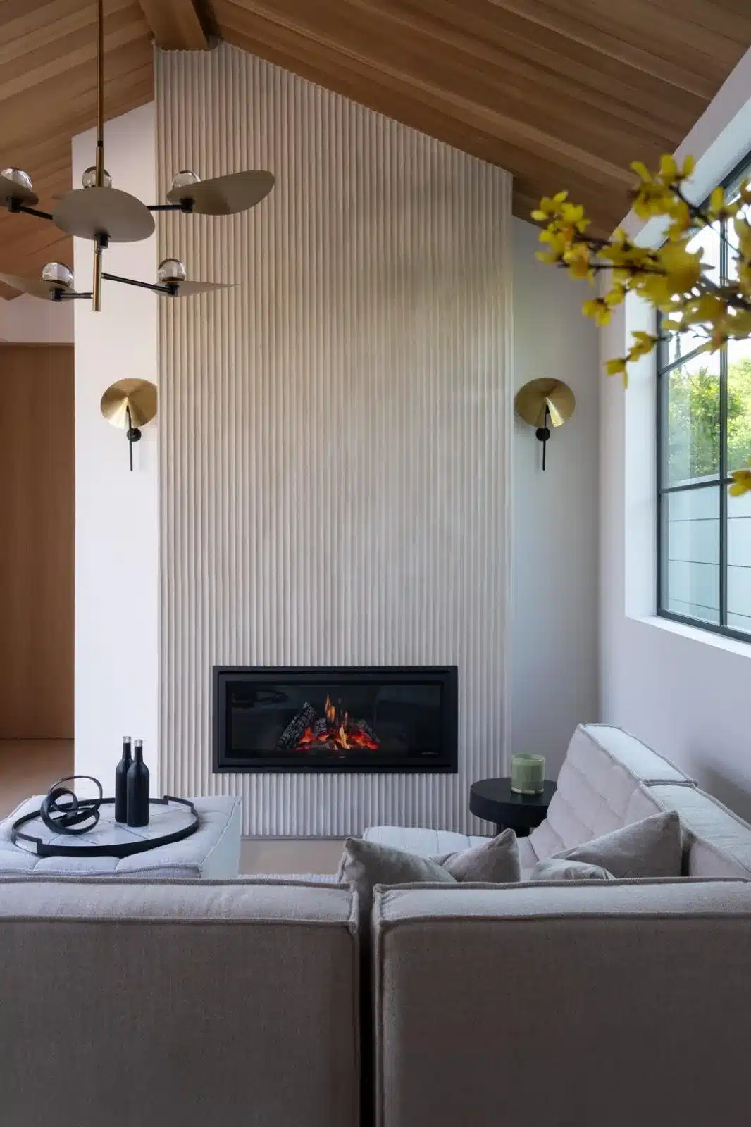Finished sitting area with a fluted white fireplace wall, sloping wood ceiling, gold-toned sconces, and gray seating