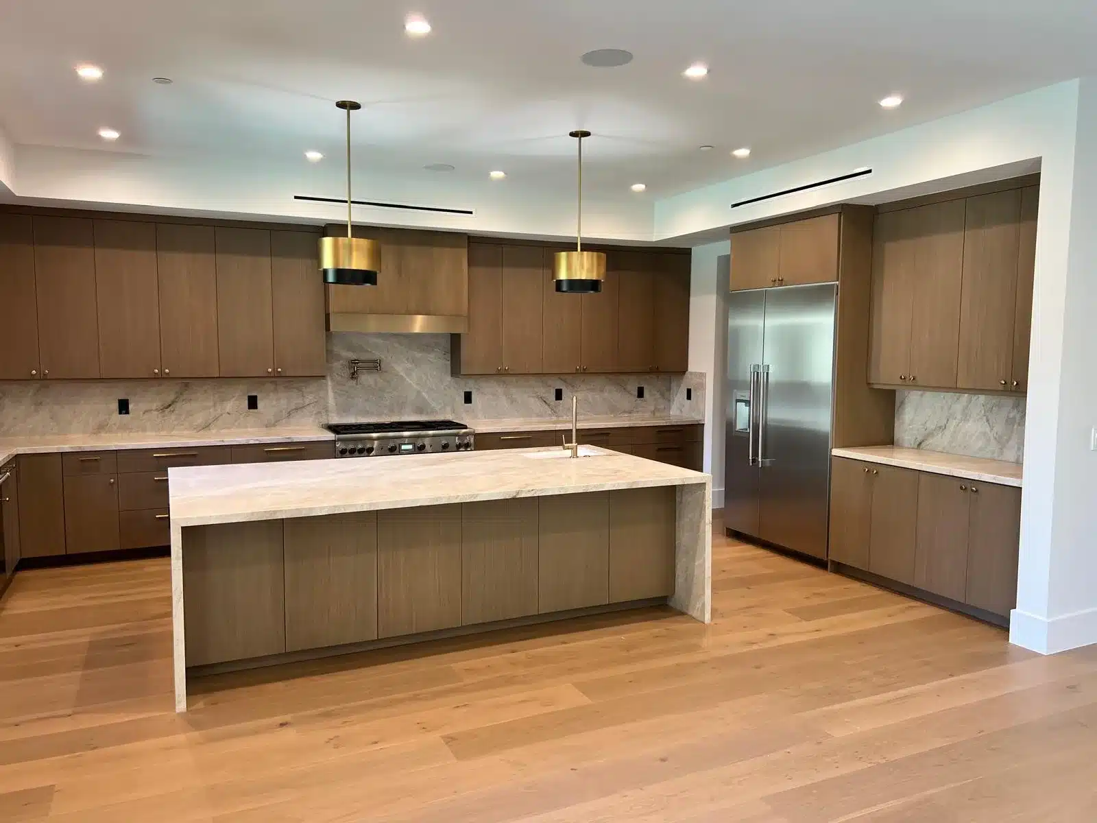 Finished kitchen viewed toward the stainless steel refrigerator and range, with wood cabinetry and a light veined island.