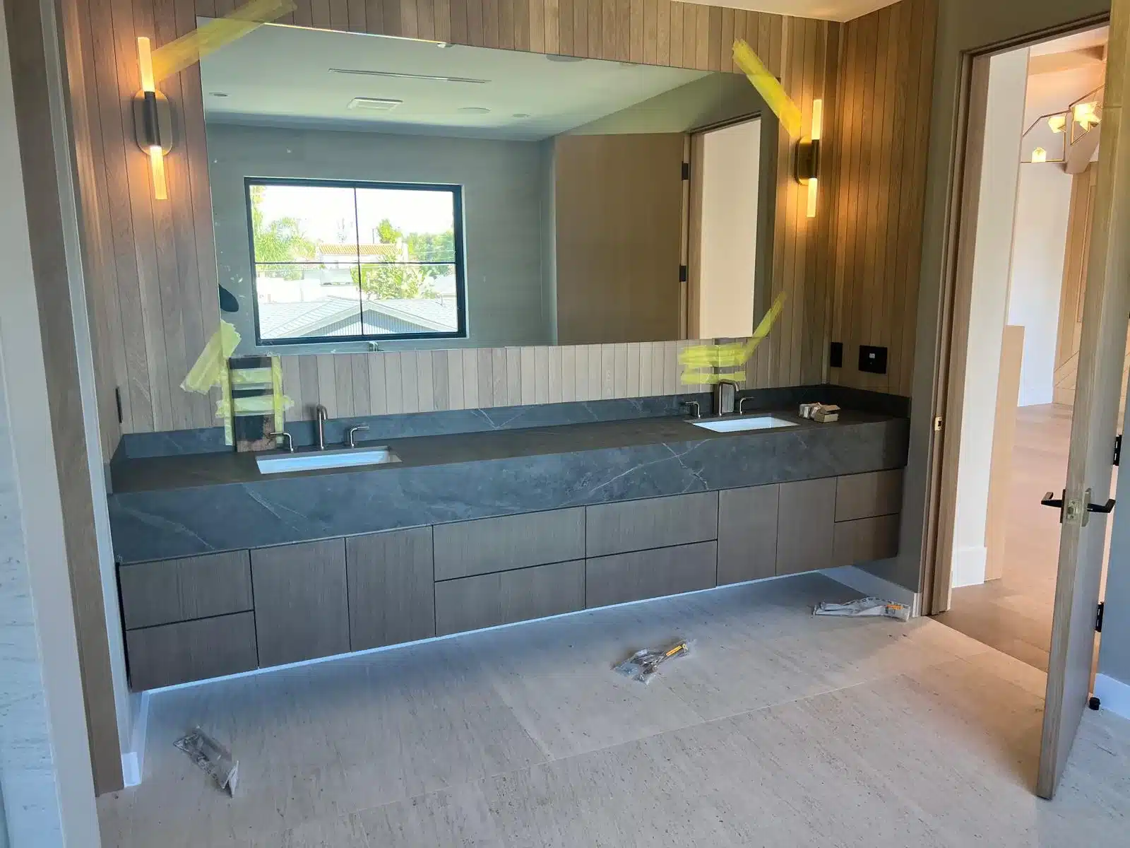 Bathroom during finishing work with a floating wood-grain vanity, dark veined countertop, inset sinks, and protective tape