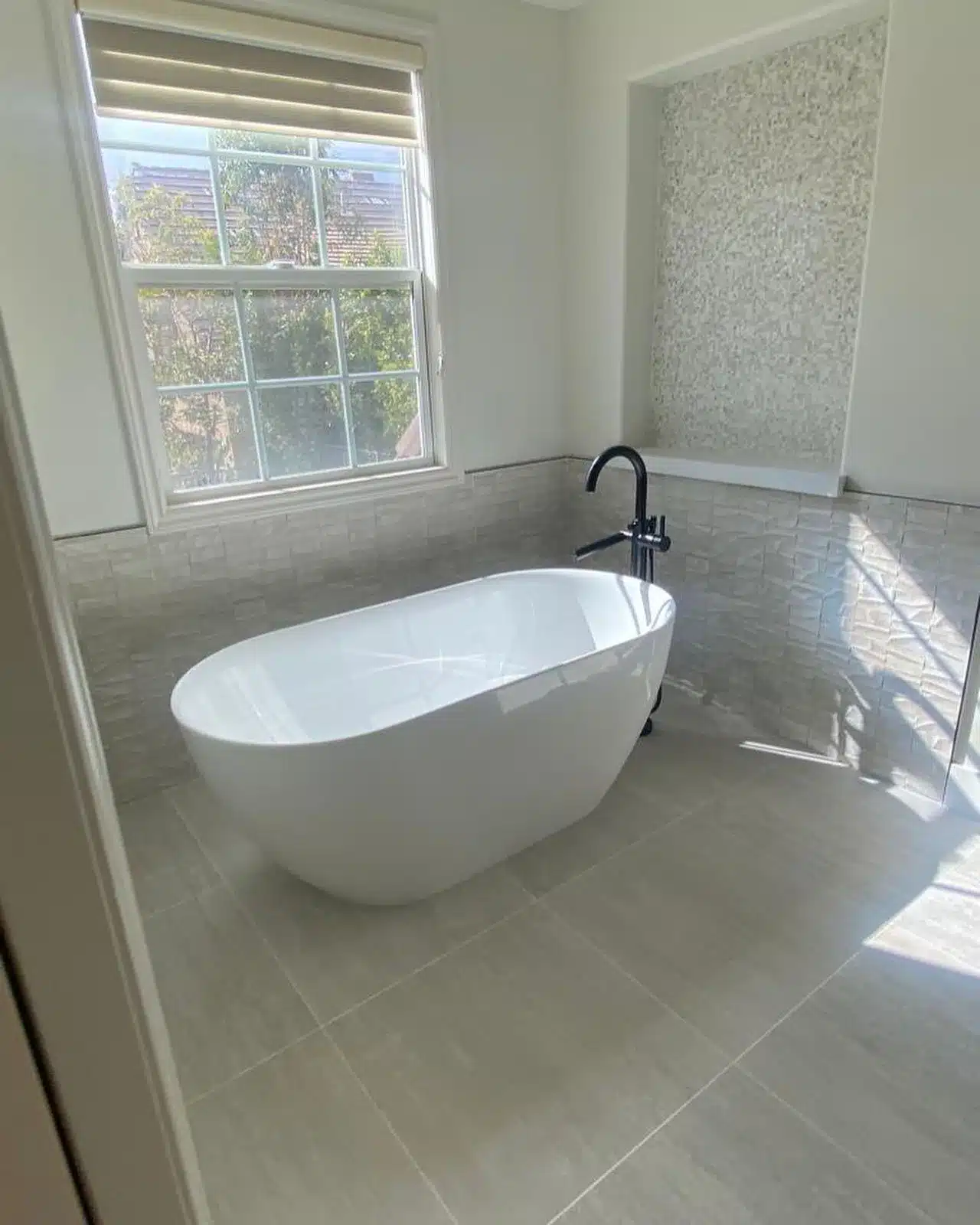 Finished bathroom with a white freestanding tub, black floor-mounted filler, gray tile, and a window with blinds.