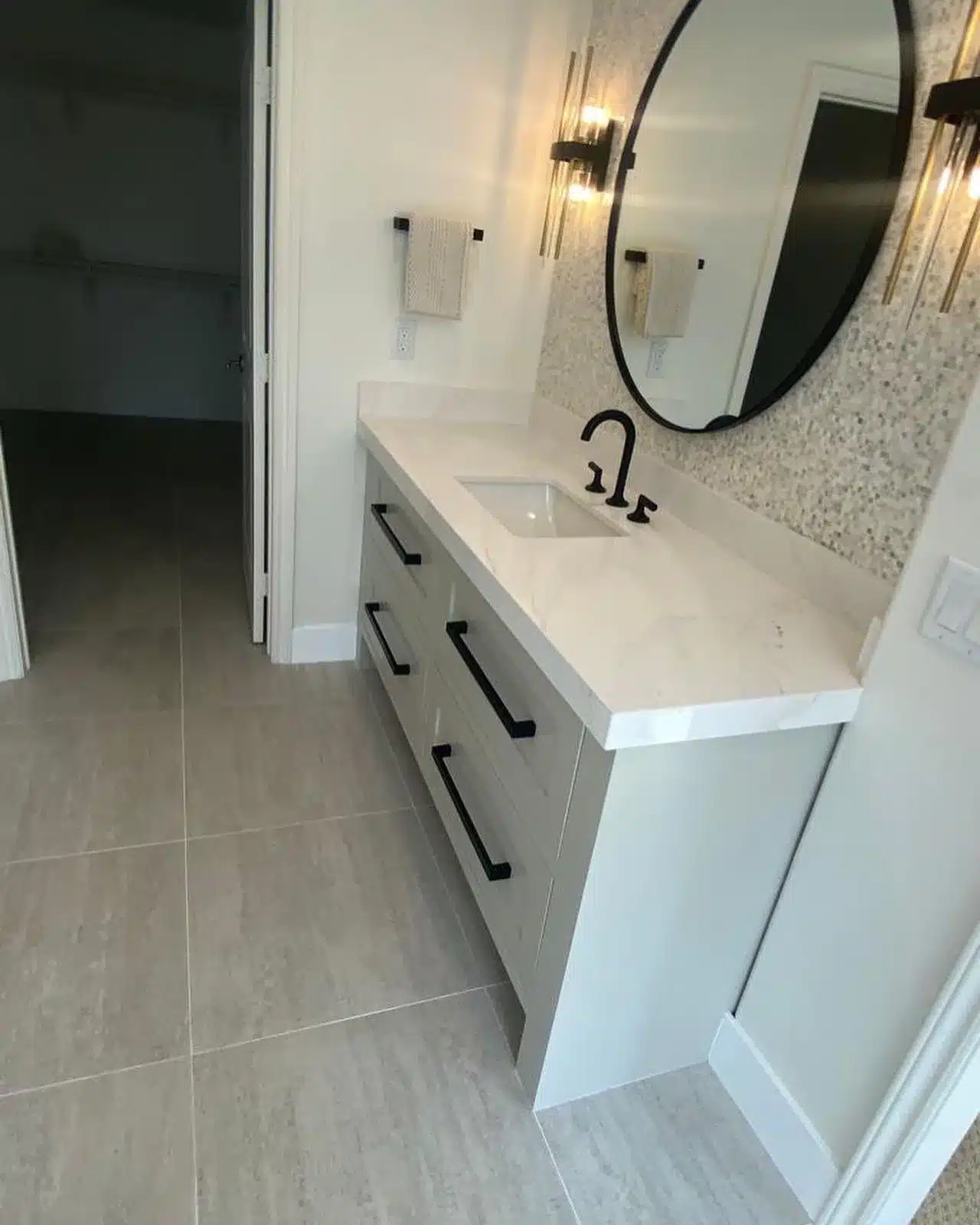 Finished light-colored vanity with black drawer pulls, a rectangular sink, round mirror, and illuminated wall sconces.