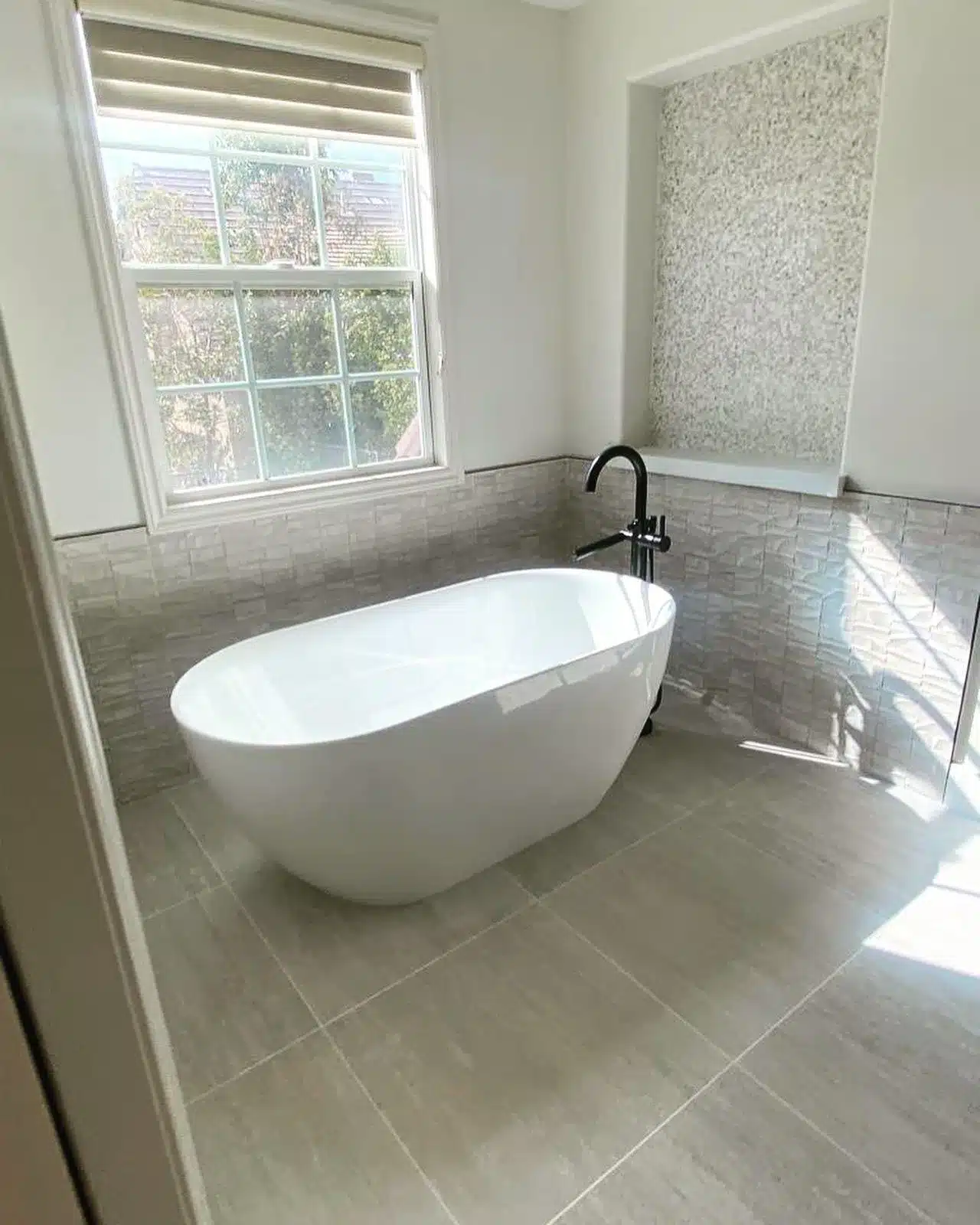 Finished white oval tub beside a window, surrounded by gray floor tile and textured wall tile.
