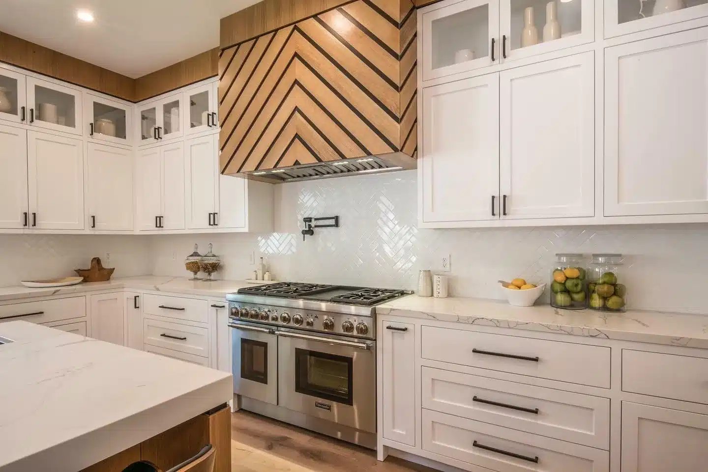 Finished range wall with a chevron-pattern wood hood, white herringbone backsplash, black cabinet pulls, and stainless steel